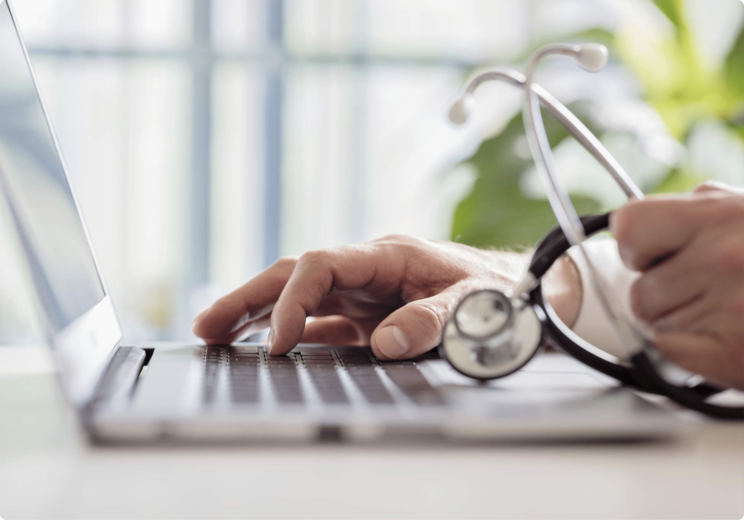 Someone typing on a laptop with a stethoscope in their other hand